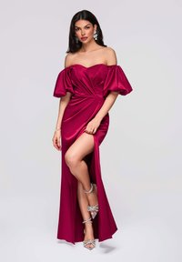 Robe en satin bordeaux avec manches bouffantes dénudant les épaules, corsage froncé, fente haute sur la cuisse et taille marquée. Complétée par des talons clairs à lanières.