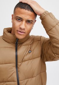 Jachetă puffer maro cu guler înalt, fermoar frontal, textură matlasată și o mică patch cu logo pe piept. Material ușor.