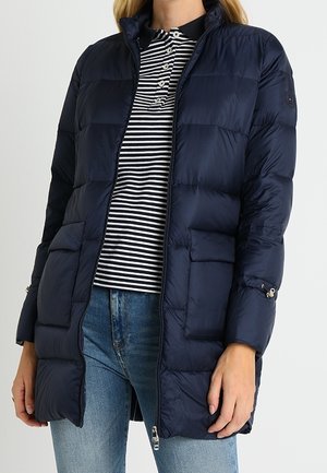 Femme portant une doudoune matelassée bleu marine sur un polo rayé noir et blanc et un jean bleu.