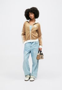 Καφέ ζακέτα με φερμουάρ και υφασμάτινα μανίκια, λευκές ρίγες, ανοιχτό μπλε τζιν παντελόνι και μια μικρή καφέ τσάντα χεριού με λογότυπο Adidas.