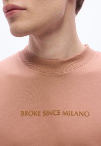 Ροζ φούτερ με ριμπ λαιμόκοψη, που φέρει τη φράση «BROKE SINCE MILANO» σε καφέ  κέντημα στο κέντρο. Απαλό, υφασμάτινο με υφή.