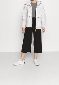 White waterproof jacket with a hood, zippered pockets, and orange accent. Paired with black culottes and white athletic shoes. Striped top beneath.