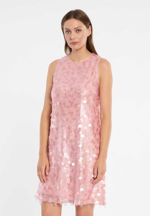 Frau in einem ärmellosen, knielangen rosa Kleid mit großen Pailletten, steht vor weißem Hintergrund, blickt nach vorne mit entspanntem Ausdruck.