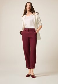 Pantaloni su misura color bordeaux con due tasche frontali e bottoni dorati in vita. Abbinati a una maglietta bianca e a un blazer color crema.