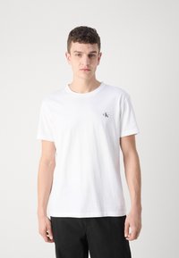 Camiseta de algodón blanca con cuello redondo y mangas cortas, con un pequeño logo negro en el lado izquierdo del pecho. Textura suave.