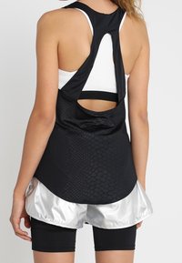 Czarny sportowy top na ramiączkach z tyłem w kształcie wyścigowym, z teksturowanym wzorem. Połączony z błyszczącymi srebrnymi szortami i czarnymi dopasowanymi legginsami.