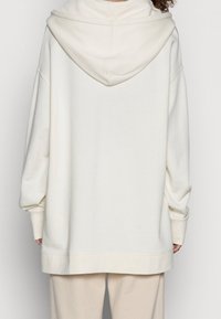 Sudadera con capucha de gran tamaño color crema, con bolsillo tipo canguro en el frente, mangas largas y capucha con cordones. Fabricada con un tejido suave y texturizado.