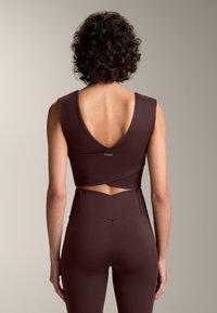 Top scurt burgundy cu un decolteu adânc la spate, textură netedă și detaliu învelit la talie, purtat cu leggings asortați cu talie înaltă.