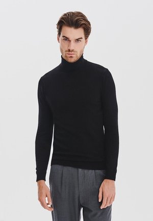 Czarny sweter z golfem, wykonany z miękkiego materiału, dopasowany krój z długimi rękawami. Model ma na sobie szare spodnie, minimalisticzne tło.