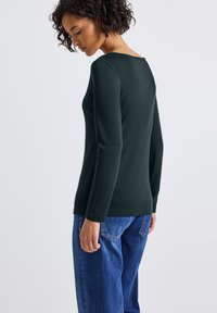 Langsleeve, donkergroen top met een wijd uitsnede. De stof lijkt zacht met minimale textuur. Gepaard met losse, vervaagde blauwe denim jeans.