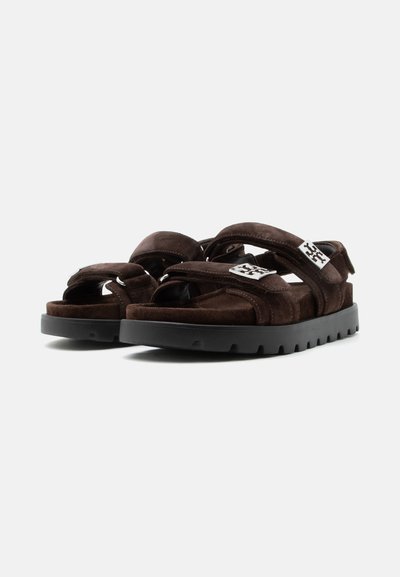 Sandales en daim brun foncé avec semelles épaisses noires et brides Velcro réglables, avec un petit logo argenté sur la bride avant.