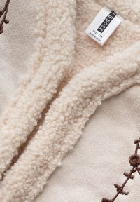 Kremowy pluszowy materiał o miękkiej, puszystej fakturze z ciemnobrązowymi haftowanymi kwiatowymi akcentami wzdłuż krawędzi.