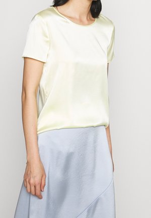 Femme portant un chemisier en satin jaune pâle à manches courtes et une jupe en satin bleu clair, debout devant un fond clair uni.