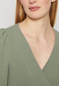 Top vert olive à col en V avec une texture légère et des détails de manches froncées, associé à des boucles d'oreilles pendantes dorées au design ondulé.