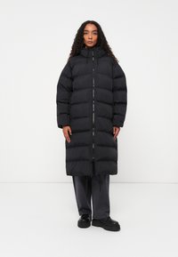 Casaco puffer preto longo com um design acolchoado, gola alta, fecho frontal e cordões ajustáveis; combinado com calças cinza escuro e botas pretas.