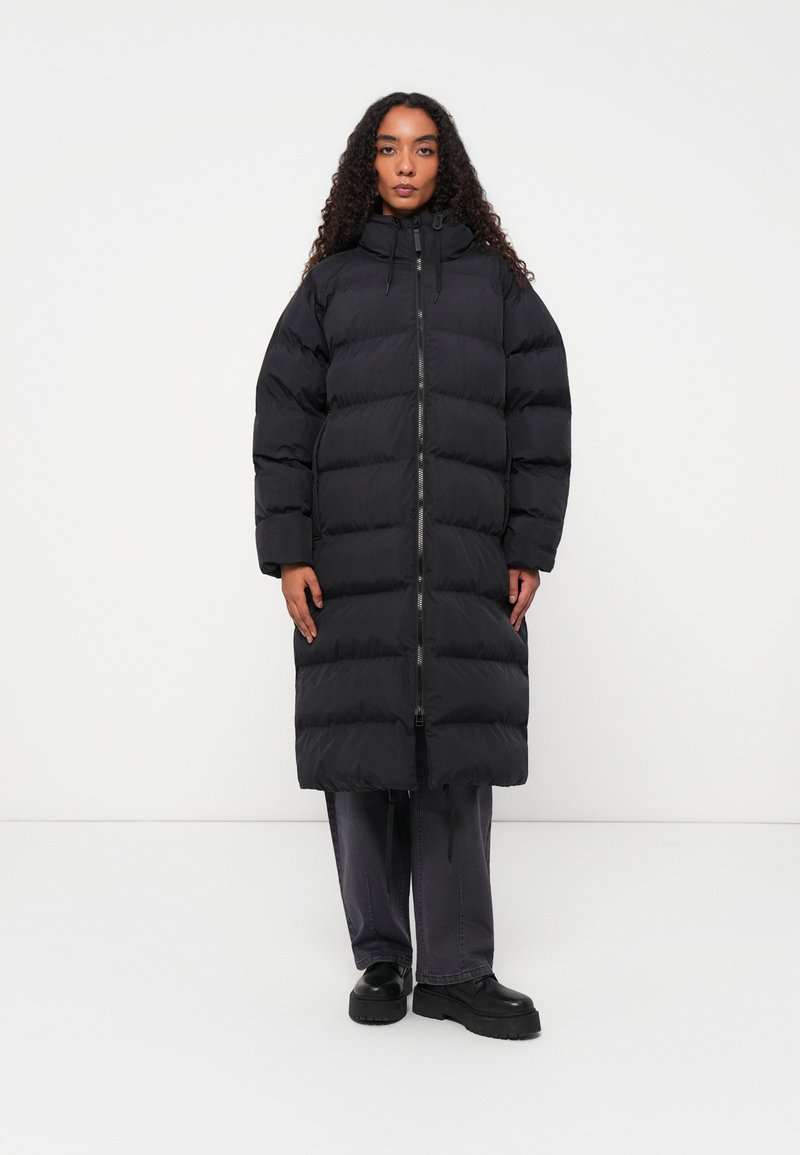 Casaco puffer preto longo com um design acolchoado, gola alta, fecho frontal e cordões ajustáveis; combinado com calças cinza escuro e botas pretas.