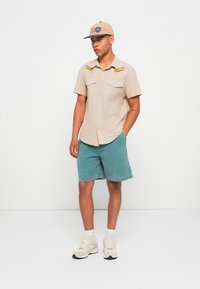 Light beige short-sleeve shirt with embroidered details, green denim shorts, and a beige cap. Footwear features a mix of cream and gray colors.