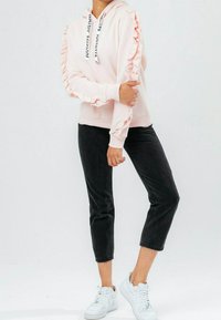 Sudadera con capucha rosa con mangas con volantes, con un cordón que lleva el texto "JUSTHYPE". Combinada con pantalones negros recortados y zapatillas blancas.