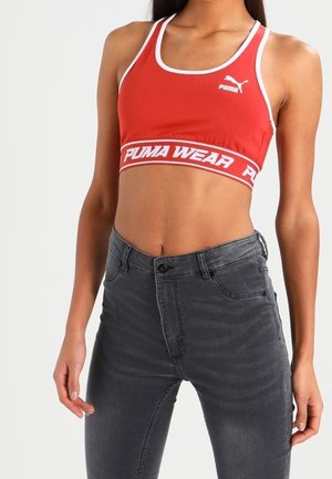 Punainen urheiluliivi valkoisilla reunoilla ja logolla, jossa on rohkea vyötärönauhateksti "PUMA WEAR." Yhdistetty tummanharmaisiin, istuviin farkkuihin.