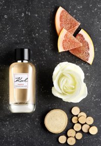 Eau de parfum parfumée dans un flacon rectangulaire transparent avec un capuchon noir. Accompagnée d'une rose blanche, de tranches de pamplemousse rose et de morceaux de bois sur une surface sombre.