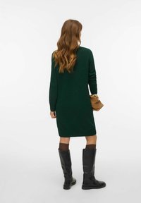 Zelené pletené šaty s dlouhými rukávy, do kolen, v kombinaci s černými kozačkami nad kolena. Vlasy volně vlnité, držící hnědou tašku.