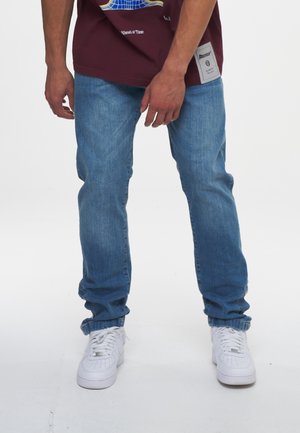 Homme portant un jean bleu clair, des baskets blanches et un t-shirt à motif bordeaux, debout devant un fond blanc uni.