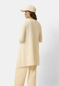 Suéter largo de manga larga beige con dobladillo acanalado y gorra beige a juego. Diseño simple, textura suave, vista desde atrás, corte relajado.