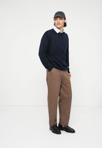 Hanorac bleumarin cu logo embossat, cămașă deschisă la culoare dedesubt, pantaloni largi maro și pantofi din piele negri. Capp cu detalii brodate.