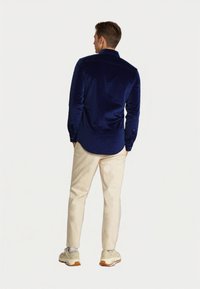 Moški v temno modri dolgem rokavu srajce, bež hlačah in belih supergah stoji s rokami v žepih, obrnjena stran.