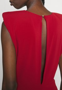 Robe rouge avec des manches courtes, présentant une découpe profonde au dos et un petit bouton au niveau du col. Texture lisse, design ajusté.