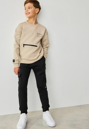 Beige sweatshirt met een ritsvak en logo, gecombineerd met zwarte cargobroek met zijzakken. Witte sneakers maken de outfit compleet.