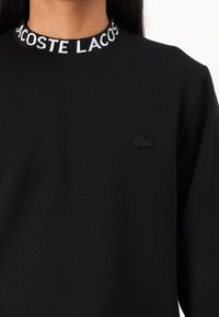 Juodas megztinis su apvaliu kaklu, puošiamas baltu "LACOSTE" logotipo juostele aplink kaklą ir mažu juodu išsiuvinėtu krokodilo logotipu ant krūtinės.