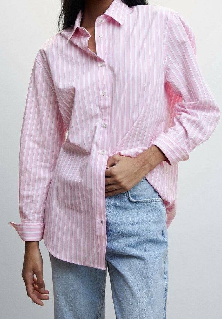 Personne portant une chemise à manches longues rayée rose et blanche à boutons, et un jean bleu clair, la main glissée dans la poche du jean.