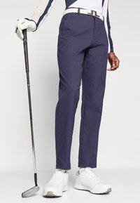 Golfhosen in Marineblau, aus leichtem Material gefertigt; verfügen über Seitentaschen, einen weißen Gürtel und sind mit weißen Sportschuhen kombiniert.