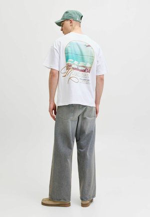 Homme portant un t-shirt blanc avec un motif de scène de plage, jean gris ample, chaussures marron clair et casquette verte, debout de dos devant un fond uni.