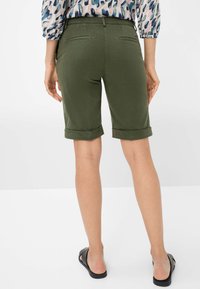 Pantaloni corti verde oliva con risvolti in tessuto morbido e tasche posteriori, abbinati a sandali neri a scorrimento, modello visto di spalle.