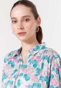 Bloemenoverhemd met een lichtgrijze achtergrond en patronen van roze en turquoise bladeren. Klassieke kraag en iets opgeblazen mouwen.