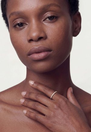 Lyhyttukkainen nainen kevyesti koskettamassa kaulaansa, sormessaan kultasormus, jossa on pieniä timantteja, neutraali tausta.