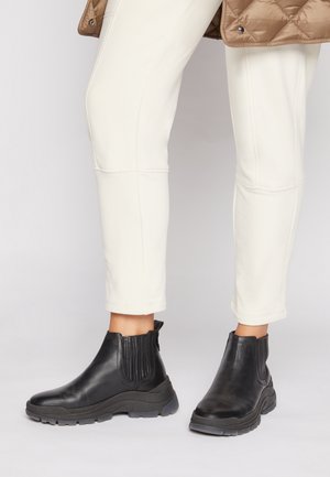 Zwarte leren enkellaarsjes met dikke zolen gedragen met gebroken witte broek en een bruine doorgestikte jas.