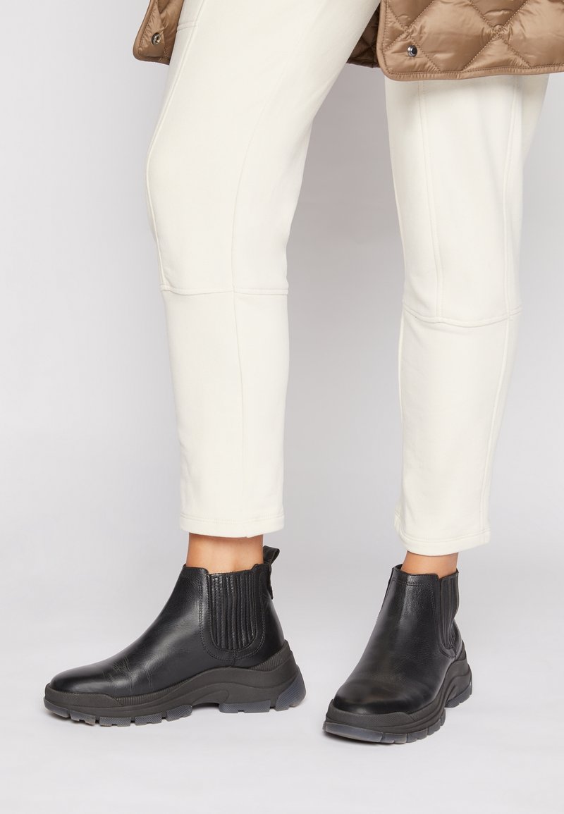 Bottines noires en cuir avec semelles épaisses portées avec un pantalon blanc cassé et une veste matelassée marron.