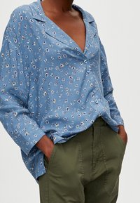 Personne portant une chemise boutonnée à fleurs bleues, ample, rentrée dans un pantalon taille haute vert olive, les mains dans les poches.