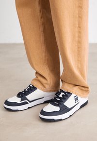 Sneakers bianchi e navy presentano una punta rotonda, un design testurizzato e un marchio distintivo. Abbinati a pantaloni tapered marrone chiaro.