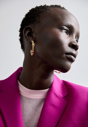Pendientes en tono dorado con acentos rojos, que presentan un diseño de cadena enlazada. El modelo lleva un blazer rosa brillante sobre una parte superior de canalé rosa claro.