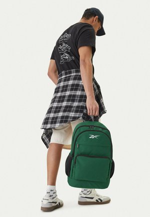 Joven con gorra negra, camiseta gráfica negra, camisa de cuadros anudada a la cintura, pantalones cortos beige, zapatillas blancas Reebok, sosteniendo una mochila verde por el asa.