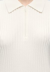 Gros plan d'un top en tricot texturé crème avec un col et une fermeture éclair verticale au niveau de l'encolure.
