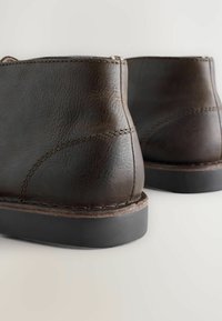 Botas de cuero marrón hasta el tobillo con una superficie texturizada, suelas de goma negras contrastantes y detalles de costura visibles a lo largo de las costuras.