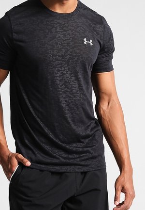 Homme portant un t-shirt noir ajusté Under Armour avec un motif camouflage subtil et un short de sport noir, debout avec une main dans la poche.
