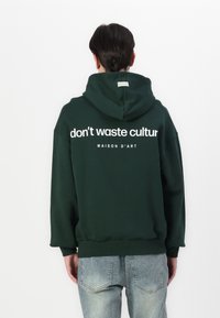 Hanorac verde cu glugă, cu text alb "nu risipi cultura" și "MAISON D'ART" imprimate pe spate; material moale, croială lejeră.