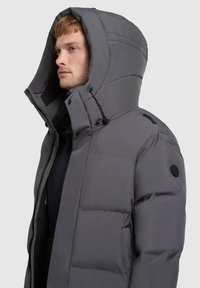 Szürke bélelt dzseki nagy kapucnival, cipzáros záródással és oldalsó zsebekkel. Kerek logófolttal és állítható húzózsinórokkal a kapucnin.