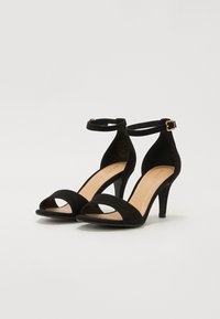 Sandalias de tacón alto de ante negro con diseño de punta abierta, tira para el tobillo y hebilla. Presentan una plantilla suave de color beige y un tacón delgado.